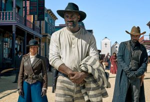 A still from the 2021 Western film The Harder They Fall showing three people walking towards us. The man in the centre is black, with a greying beard, and wears a ten-gallon black hat, a white long-sleeved vest and striped prison trousers, with the striped jacket of the uniform over his arm. To the right, another black man in a cowboy hat and long coat; to the left, a black woman in leather jacket and black hat.